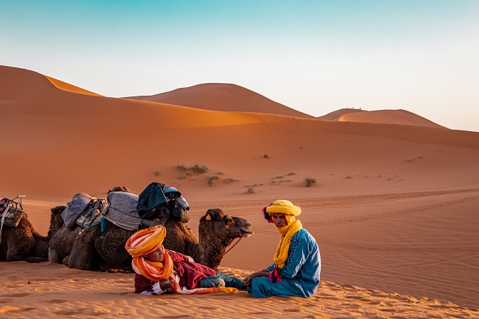 Il deserto del Marocco è un’esperienza diversa a seconda della regione che si sceglie per visitarlo: nella foto le dune sahariana di Merzouga, con due esemplari di cammello e un berbero seduti.